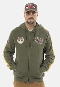 Sweat à capuche zippé olive avec des rayures jaunes sur les côtés, arborant des patchs Kustom et Von Dutch. Fabriqué en tissu doux avec une poche avant.