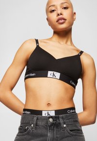 Svart bomullsbH med V-halsdesign og justerbare stropper, med et merket elastisk bånd som leser "Calvin Klein" i hvit tekst.