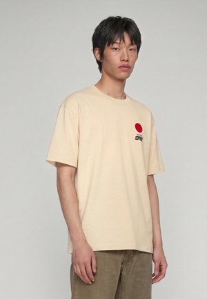 Ung mand med kort sort hår iført en beige T-shirt med en rød cirkel og sort tekst på brystet, står mod en ensfarvet lys baggrund.