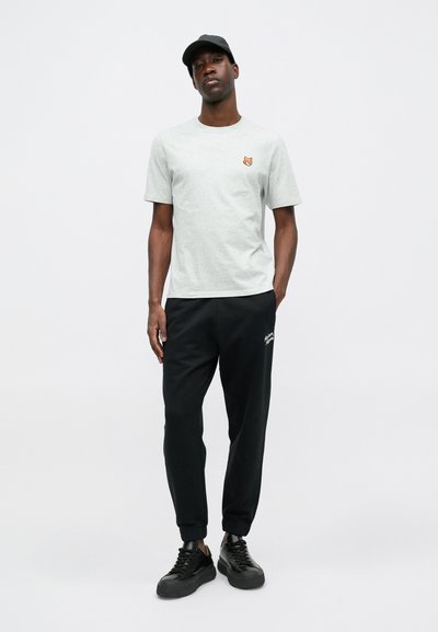 Tricou gri cu un mic logo portocaliu, asociat cu pantaloni sport negri care au text alb. Purtați cu o șapcă neagră și adidași negri.