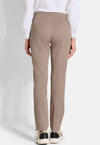 Vrouw in beige rechte broek, witte blouse met lange mouwen en beige sneakers met zwarte accenten, staande tegen een effen achtergrond.