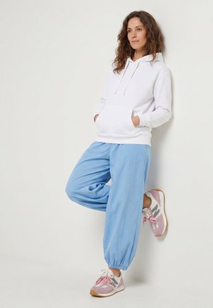 Frau lehnt an einer Wand, trägt einen weißen Hoodie, hellblaue Hose und rosa-weiße Sneaker, die Hände in den Taschen, schaut nach vorne.
