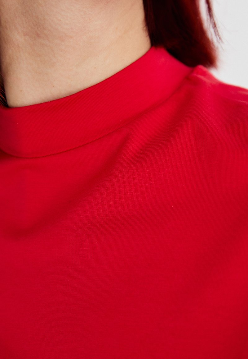 Czerwona bluzka z wysokim dekoltem, wykonana z gładkiego materiału, charakteryzująca się dopasowanym krojem oraz czystą, minimalistyczną estetyką bez widocznych wzorów czy ozdób.