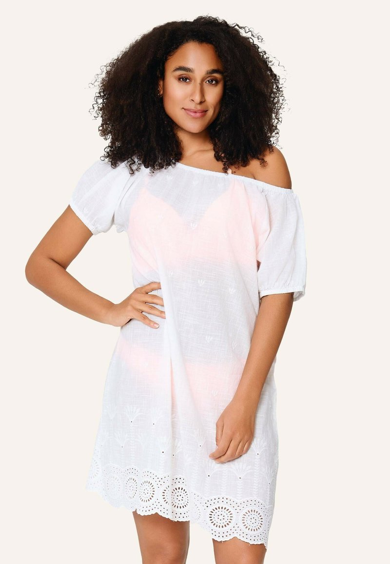 Žena s kučeravými vlasmi v bielych šatách bez ramienok s čipkovaným lemom, stojaca s rukou na boku pred jednoliatym pozadím.