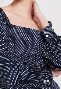 Blusa azul marino con lunares blancos. Presenta un escote cuadrado y mangas abullonadas. Termina con botones en forma de corazón en los puños.