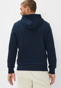 Felpa con cappuccio blu navy realizzata in materiale morbido, con cappuccio regolabile con coulisse, polsini e orlo a coste, caratterizzata da un design semplice, indossata sopra pantaloni chiari.