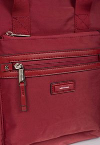 Zaino di tessuto rosso con superficie testurizzata, dotato di tasca con zip, etichetta con marchio e punti di attacco con cinturini rinforzati.