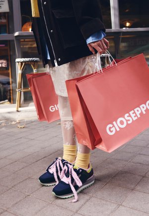 Bolsas de compra rojas con el logo "Gioseppo" en blanco, sujetas junto a medias con estampado y zapatillas náuticas azul marino con cordones rosas y detalles verdes.
