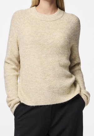 Beige trui met geribbelde stof en lange mouwen, gedragen door een persoon met de handen in de zakken van een zwarte broek tegen een witte achtergrond.