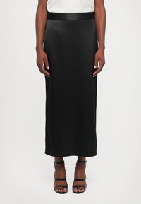 Vrouw in een zwarte midi pencil skirt met een satijnen tailleband en zwarte open teen enkelband hakken, staand tegen een witte achtergrond.