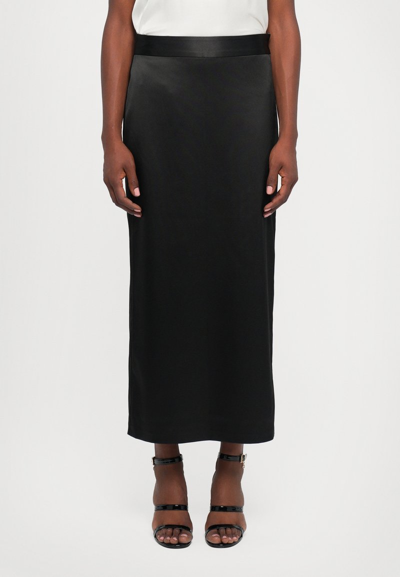Vrouw in een zwarte midi pencil skirt met een satijnen tailleband en zwarte open teen enkelband hakken, staand tegen een witte achtergrond.