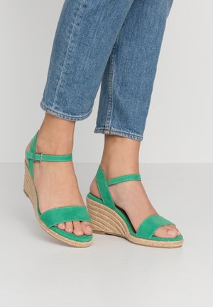 Fødder iført grønne kileheels-sandaler med ankelremme, kombineret med blå korte jeans på en ensfarvet lys baggrund.