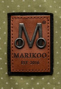 Brown leather label with embossed silver "M" and black accents, stitched onto olive green fabric with white triangular patterns.