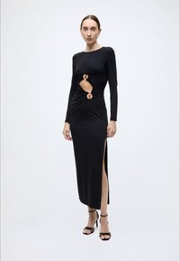 Zwarte jurk met lange mouwen, voorzijde geknoopt, met uitsparingen en cirkelvormige accenten. Heeft een zijsplit. Gemaakt van glad, rekbaar materiaal.