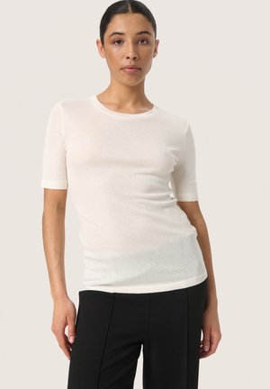 Vit kortärmad t-shirt tillverkad av lätt, genomskinlig tyg. Har rund halsringning och en figurnära design, tillsammans med svarta byxor.