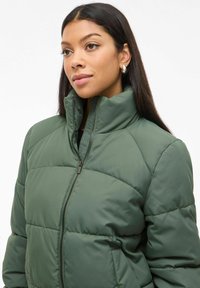 Grøn quilted puffer jakke med høj krave og lynlås. Har en glat tekstur og vandret syninger for ekstra isolering.