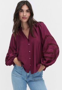 Frau in einer lockeren, langärmligen, knopfbaren lila Bluse mit Spitzen-Details und hellblauen Jeans, Hände in den Taschen, neutraler Hintergrund.