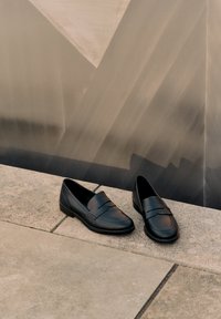 Par sorte læder loafers placeret side om side på en udendørs stensurface med en metallisk væg i baggrunden.