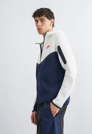 Młody mężczyzna w białej i granatowej kurtce na zamek marki Nike z czerwonym logo, czarne i granatowe spodnie, ręka w kieszeni, zwrócony lekko w lewo.