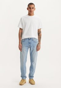 Witte katoenen t-shirt, lichtblauwe denim jeans, beige moccasin schoenen. Persoon met tatoeages staat tegen een neutrale achtergrond.