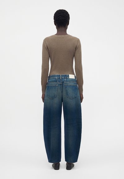 Personne aux cheveux courts et bouclés portant un haut marron à manches longues et un jean bleu à jambes larges, debout de dos sur fond blanc.