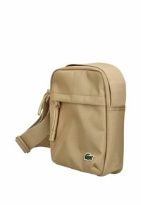 Borsa rettangolare marrone chiaro con superficie testurizzata, tracolla regolabile e chiusura a zip, dotata di un piccolo logo verde Lacoste.