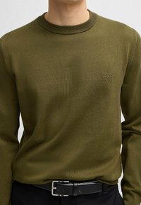Oliwkowy sweter dzianinowy z okrągłym dekoltem i ściągaczem. Posiada subtelne logo "BOSS" wyszyte na klatce piersiowej. Noszony na ciemnych spodniach i czarnym pasku.
