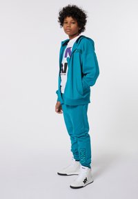 Haut à capuche teal et jogging assorti en tissu doux. T-shirt graphique blanc en dessous, accompagné de baskets montantes blanches avec des accents noirs.