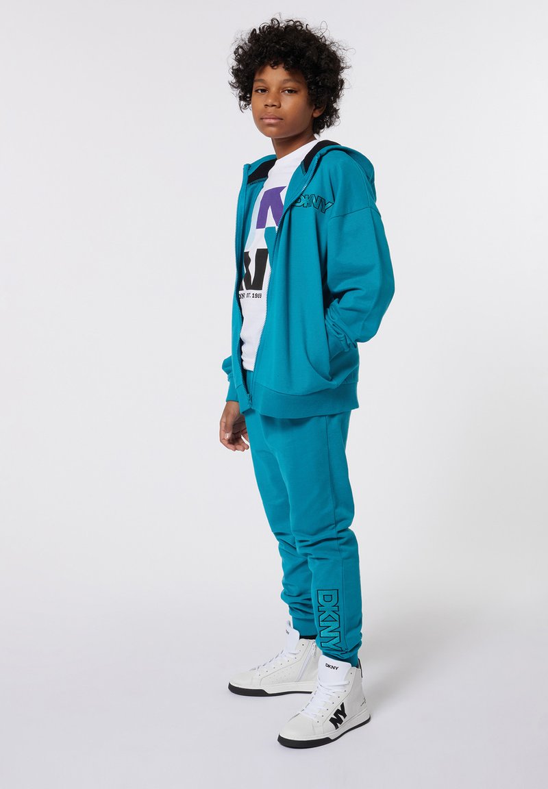 Haut à capuche teal et jogging assorti en tissu doux. T-shirt graphique blanc en dessous, accompagné de baskets montantes blanches avec des accents noirs.