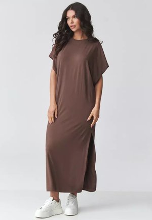 Kobieta o długich, ciemnych, kręconych włosach, ubrana w luźną, brązową sukienkę maxi z bocznym rozcięciem i białe trampki na platformie, stoi na tle jednolitego tła.