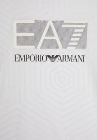 Camiseta blanca con un patrón geométrico negro y el texto "E A" y "EMPORIO ARMANI" en una fuente negrita. Tejido suave, diseño minimalista.