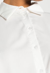 Bílá blůza s klasickým límečkem, zapínáním na knoflíky, hladkou texturou a průhlednými knoflíky. Detaily podtrhují čistý, minimalistický design.