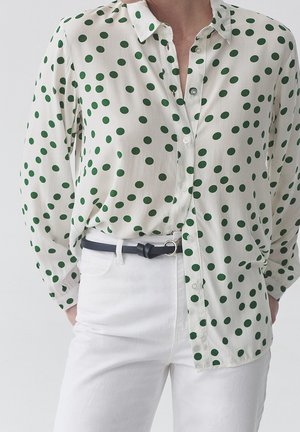 Femme portant un chemisier blanc à boutons avec des pois verts, rentré dans un pantalon blanc taille haute avec une ceinture noire.