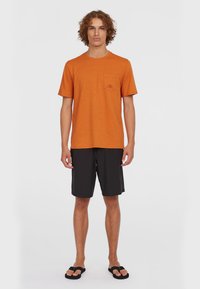 Orange kortærmet t-shirt med en brystlomme og logo, parret med sorte shorts og sorte klip-klapper, stående foran en ensfarvet baggrund.