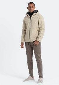 Beige vattentät jacka med huva, dragkedjeförsedd framficka, elastiska ärmslut, bärs över en svart turtleneck och bruna byxor med grå sneakers.