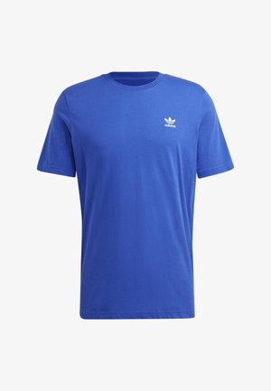 Man draagt een blauw Adidas T-shirt en zwarte Adidas trainingsbroek met logo's, staand met een hand in de zak tegen een witte achtergrond.