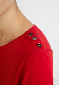 Roter Pullover mit gerippter Textur, der drei dekorative schwarze Knöpfe mit roten Akzenten an der linken Schulter aufweist.