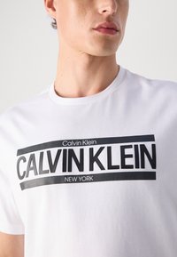 White cotton t-shirt featuring bold black text "CALVIN KLEIN" and "NEW YORK" in a rectangular design on the front. Short sleeves and crew neck.