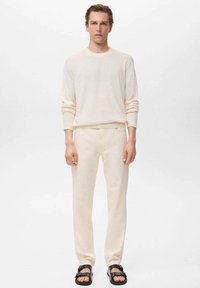 Maglione beige chiaro a maniche lunghe abbinato a pantaloni coordinati, realizzato in tessuto morbido, con un taglio rilassato e design minimal. Sandali neri completano l'outfit.