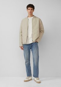 Gequilted beige bomberjack, witte henley shirt, blauwe spijkerbroek en beige sneakers. Bevat een ritssluiting en geribbelde manchetten.