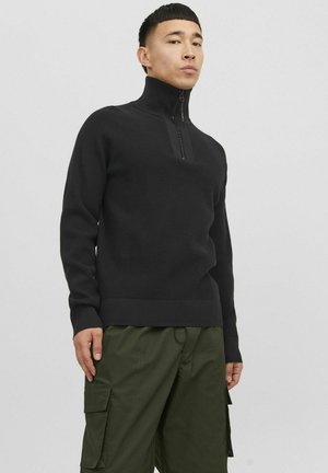 Jersey negro de punto con cuello alto y cremallera corta en la parte delantera. Puños y dobladillo acanalados. Combinado con pantalones cargo verdes que cuentan con bolsillos laterales.