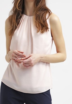 Femme portant un chemisier sans manches rose clair avec des plis près de l'encolure et un pantalon foncé, les mains légèrement jointes à la taille.