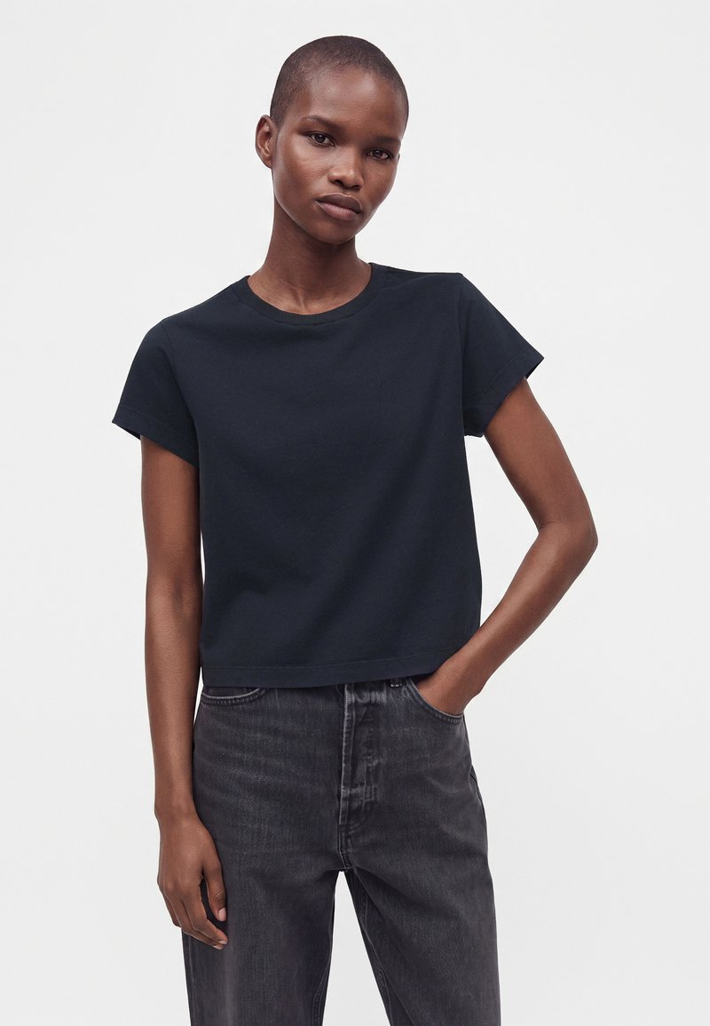 Personne aux cheveux courts portant un t-shirt noir uni à manches courtes et un jean délavé foncé, debout avec une main dans la poche.