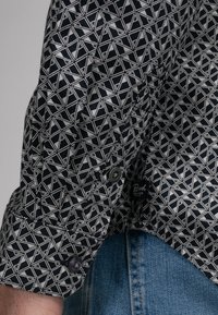 Czarna i biała koszula w geometryczne wzory, z mankietami na guziki i teksturowanym materiałem, w połączeniu z niebieskimi dżinsami.