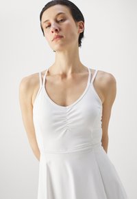 White dress with a fitted bodice, thin straps, and a gathered detail at the center. Flared skirt with a textured waistband. Smooth fabric.