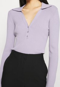 Fialový žebrovaný pletený top s výstřihem do V a knoflíkovým zapínáním, s klasickým límcem a dlouhými rukávy, doplněný černými kalhotami.