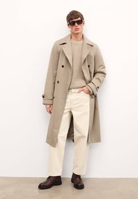 Trench coat bej cu nasturi dubli, pulover bej cu dungi, pantaloni crem și cizme maro cu șireturi, completate cu ochelari de soare negri.