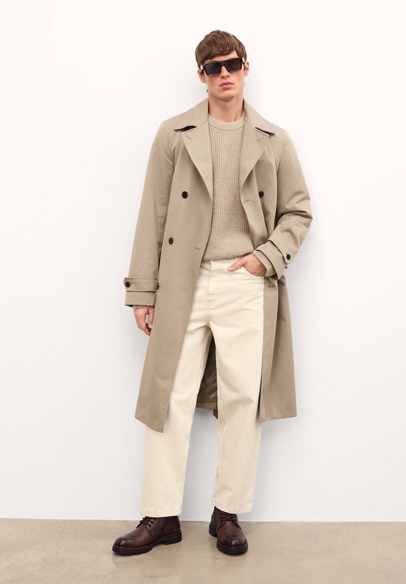 Trench coat bej cu nasturi dubli, pulover bej cu dungi, pantaloni crem și cizme maro cu șireturi, completate cu ochelari de soare negri.
