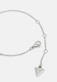 Silberne Kettenarmband mit einem Karabinerverschluss und einem dreieckigen Anhänger, der das eingravierte Wort "GUESS" und ein Fragezeichen zeigt.