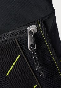 Svart ryggsäck med ett texturerat nätpanel, silverfärgade dragkedjefästen och klara gula detaljer. Materialet har en tålig, slät yta.